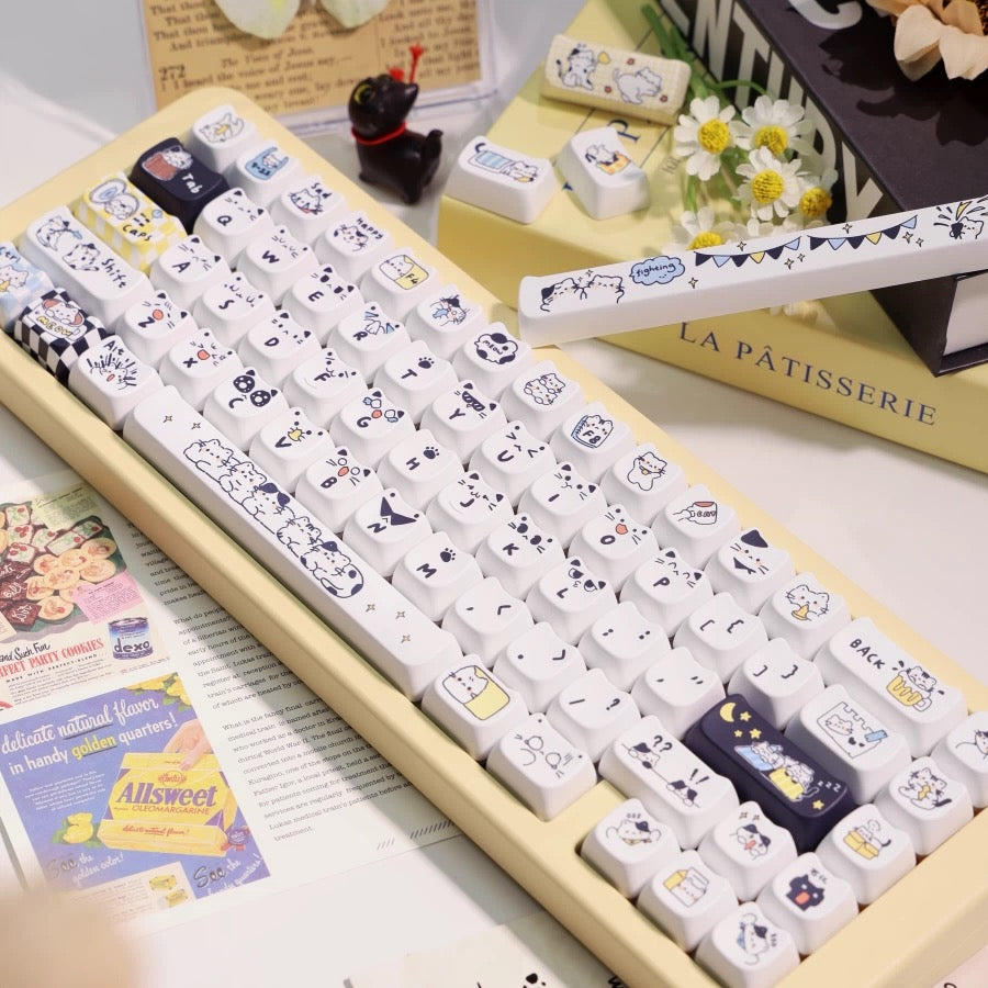 Cat-themed keycaps on white keys with black accents, laid out in cream display tray