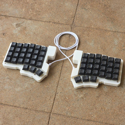 Split wired keyboard with dark keycaps, light base, white cable, angled pair.
